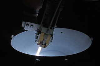 A robotic machining head operates on a white circular disk in a dark environment.