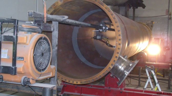 A large hollow cylindrical metal pipe with a flange sits on a red support frame in an industrial workshop, surrounded by machinery.