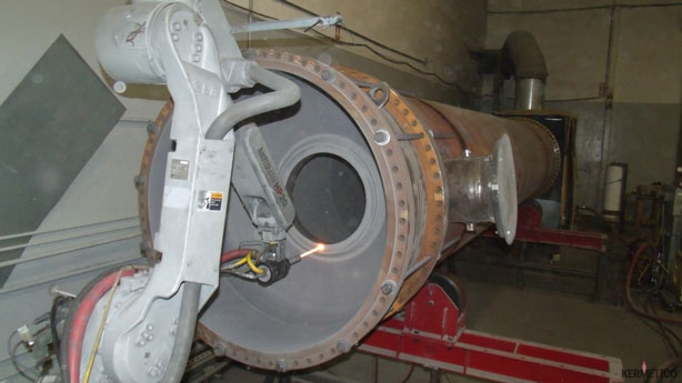 Large cylindrical metal vacuum chamber with a bolted circular flange mounted on a red stand in a workshop.