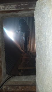 A dark, narrow industrial shaft with a bright beam of light from the left, corrugated metal lining on one side and a rough concrete wall on the other, with a vertical structure running down the center.