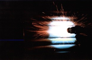 Silhouette of a person welding in the dark, with a bright arc and shower of orange sparks.