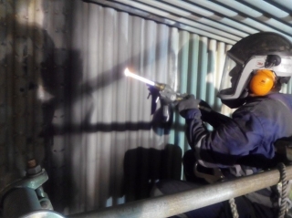 A worker wearing a welding helmet and protective gear welds a metal panel, with a bright torch flame visible.