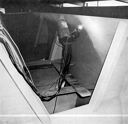 A worker in protective gear welds inside a metal framework, sparks flying from a handheld torch.
