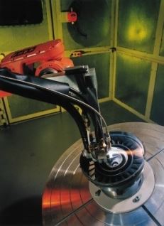 Close-up of a shiny metal circular disk with a red handheld tool and hoses against yellow-green cabinet panels.