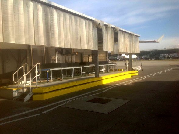 An airport loading dock with a metal canopy, stairs on the left, a bright yellow safety curb along the edge, and a distant airplane on the tarmac.