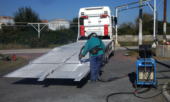 Worker in a teal shirt guides a metal loading ramp extended from a white truck, with a blue generator nearby.