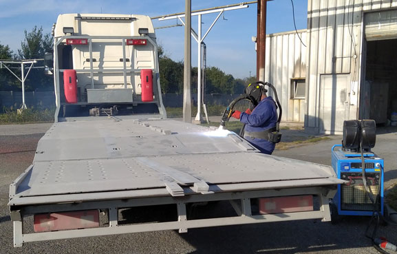 A worker in blue overalls crouches beside a white flatbed truck trailer in an industrial yard, with a blue welding machine nearby.