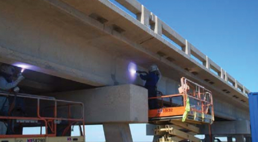 Two workers on a raised orange manlift perform maintenance on a concrete bridge support beneath a clear blue sky.