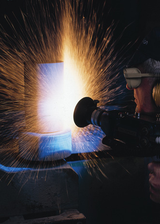 A welder in protective gear uses a torch on a glowing blue-hot piece of metal, with a shower of sparks flying.