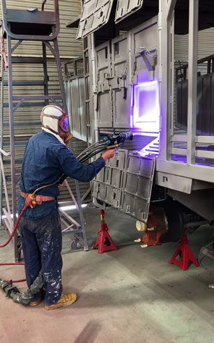 A welder in protective gear welds a metal panel on a train car inside a workshop, with a bright purple welding arc.