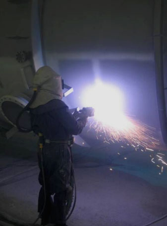 A worker in a heavy protective welding suit welds metal, sparks flying from the arc.
