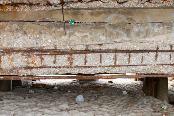 Rusty perforated metal beam under a weathered concrete surface, with a sandy, rubble-strewn ground beneath.