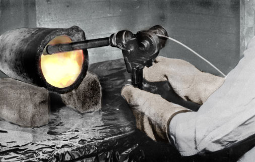 A glassblower heats glass in a glowing furnace with a handheld torch.