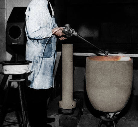 A worker in protective clothing and gloves uses tongs to handle molten material in a large crucible beside a furnace.