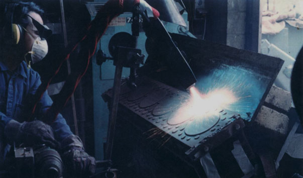 A worker in protective gear welds a metal box in a dark workshop, with a bright arc and sparks.