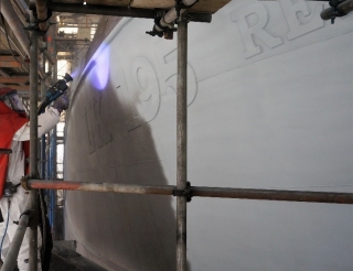 A construction worker in an orange safety vest stands beside a large white wall panel mounted on scaffolding.