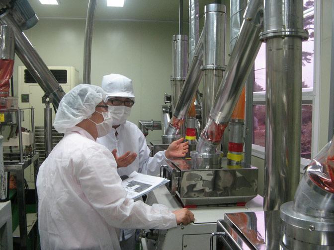 Two workers in white cleanroom outfits with hairnets and masks operate stainless steel production equipment.