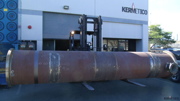 A large, rusty cylindrical metal tank lying on its side in a parking lot outside an industrial building.