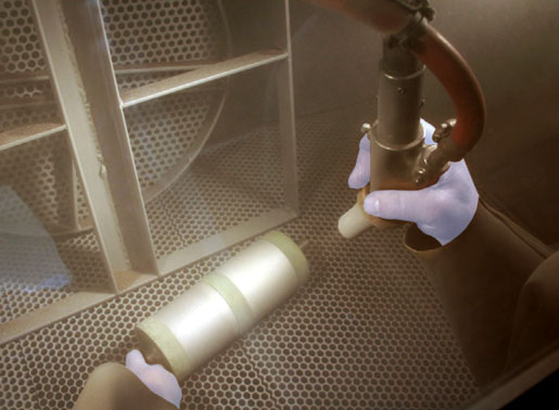 Gloved hands hold a spray bottle and a curved hose nozzle inside a stainless steel, perforated metal workspace.