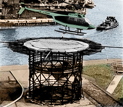 A green helicopter hovers above a circular metal tower by the water, with a dock and boat visible in the background.
