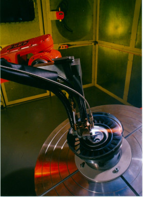 Close-up of a turntable tonearm over a shiny vinyl record in a metallic, industrial-looking enclosure.