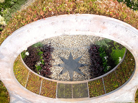 Overhead view of a circular garden with a star-shaped central design, surrounded by concentric ring walkways and a railing.