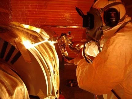 Technician in a protective suit and gas mask works on a large circular turbine in an orange-lit industrial setting.