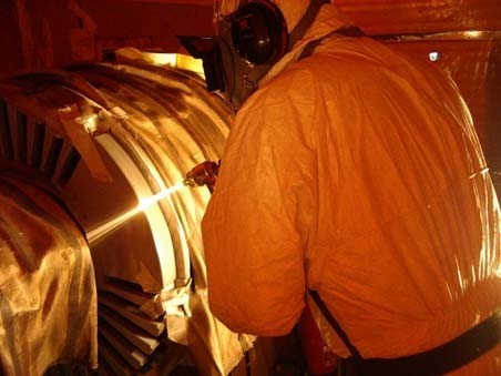 A worker in an orange protective suit and helmet inspects a large industrial turbine with visible rotor blades.