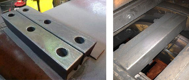 Two metal components in a workshop: a perforated clamp block with holes and a long steel beam resting on a track.