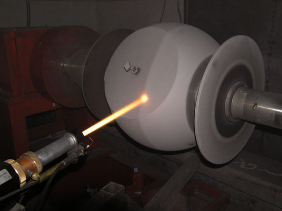 A white spherical object is mounted on a grinding spindle while an orange torch flame is directed at its surface in a workshop.