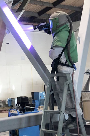 Person wearing a green protective suit and face shield stands on a ladder, welding a metal beam with a purple arc in a workshop.