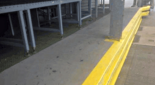 Concrete sidewalk with a bright yellow safety line along its edge beside a building and metal posts.