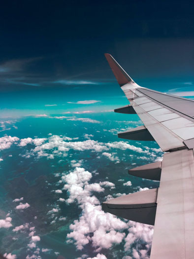 Aerial view of an airplane wing over white clouds and a turquoise sea.