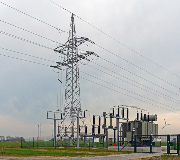 Electrical substation with lattice transmission towers and power lines in a rural landscape, with a distant wind turbine on the horizon.