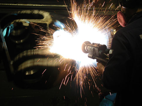 A person wearing protective gear uses an angle grinder, producing a shower of sparks beside a car.