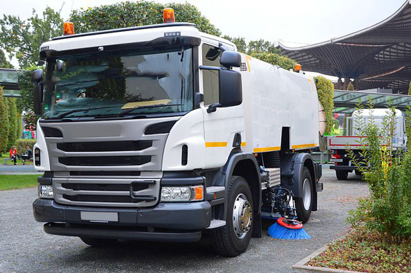White street-sweeping truck with orange beacon lights and a blue side brush, parked on a paved lot.