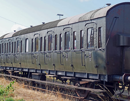An old green passenger train carriage on a railway track beside dry grass under a blue sky.