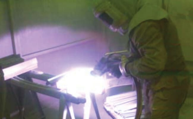 A person in protective clothing and gloves welds at a metal workbench, with a bright welding arc.