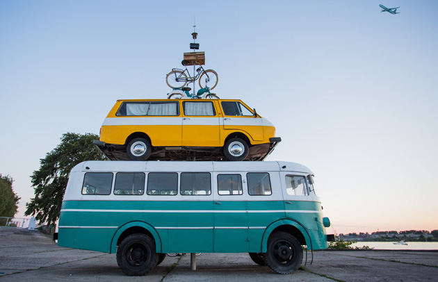 A yellow vintage van perched atop a turquoise bus, with a bicycle on the van’s roof, by the water at sunset.
