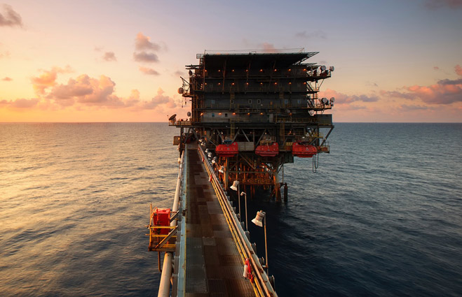 An offshore platform connected by a long walkway over the ocean at sunset, with red lifeboats on the structure.