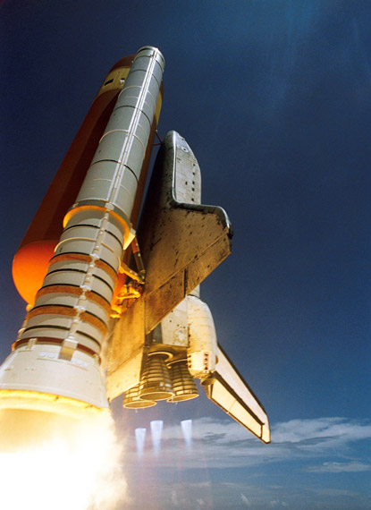 Space shuttle lifting off with an orange external fuel tank and boosters, engines blazing against a blue sky.