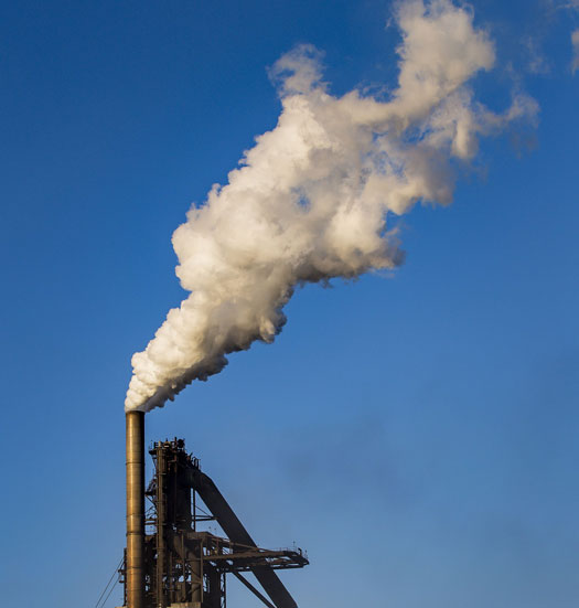 Tall industrial smokestack with a metal framework releases a plume of white smoke into a clear blue sky.
