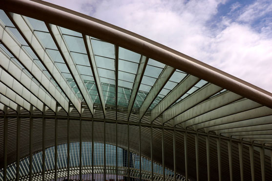 Low-angle view of a curved glass roof canopy with metal ribs and a lattice railing, set against a partly cloudy sky.