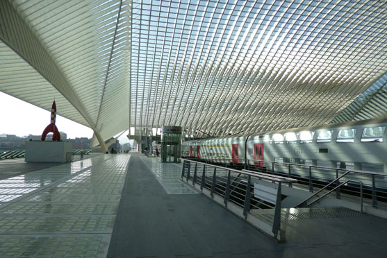 A long, bright pedestrian concourse under a grid-pattern curved roof with glass walls, railings, and a distant red sculpture.