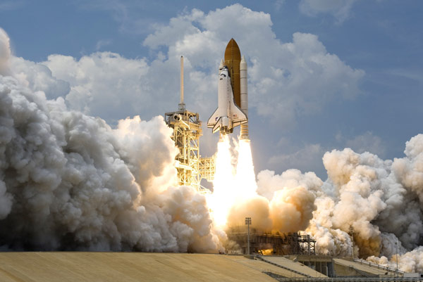 A space shuttle lifts off from a launch pad, surrounded by a massive plume of smoke and flames against a blue sky.