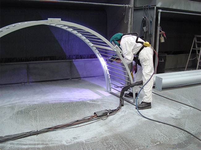 A worker in a white hazmat suit and green hood uses a tool beside a curved metal arch in an industrial workspace.