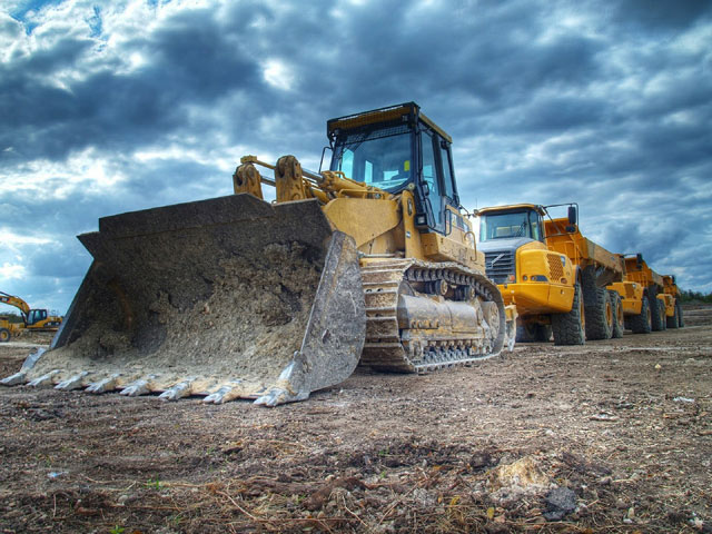 A yellow bulldozer with a large front blade sits on a dirt lot beneath a cloudy sky, with another yellow construction vehicle in the background.