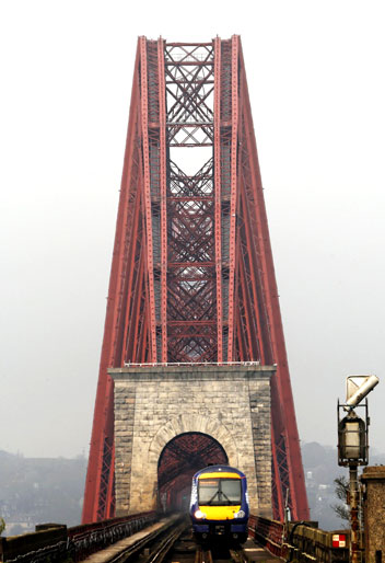 Red steel lattice bridge towering over a stone approach, with a blue and yellow train on the tracks in the foreground.