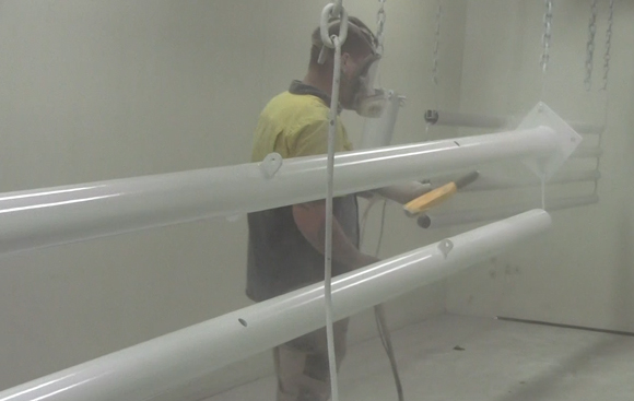 A worker wearing a yellow shirt and a respirator stands in a spray booth beside large white cylindrical pipes suspended from the ceiling.