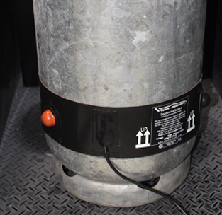 Close-up of a galvanized metal cylinder with a black band, orange button, and warning label on a diamond-plate floor.